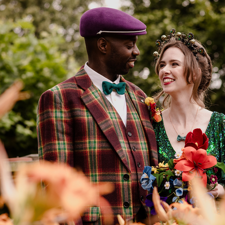 A wedding groom in a green bow tie and burgundy vintage style suit smiles at his bride in a green sparkly dress and matching mini bow tie necklace.