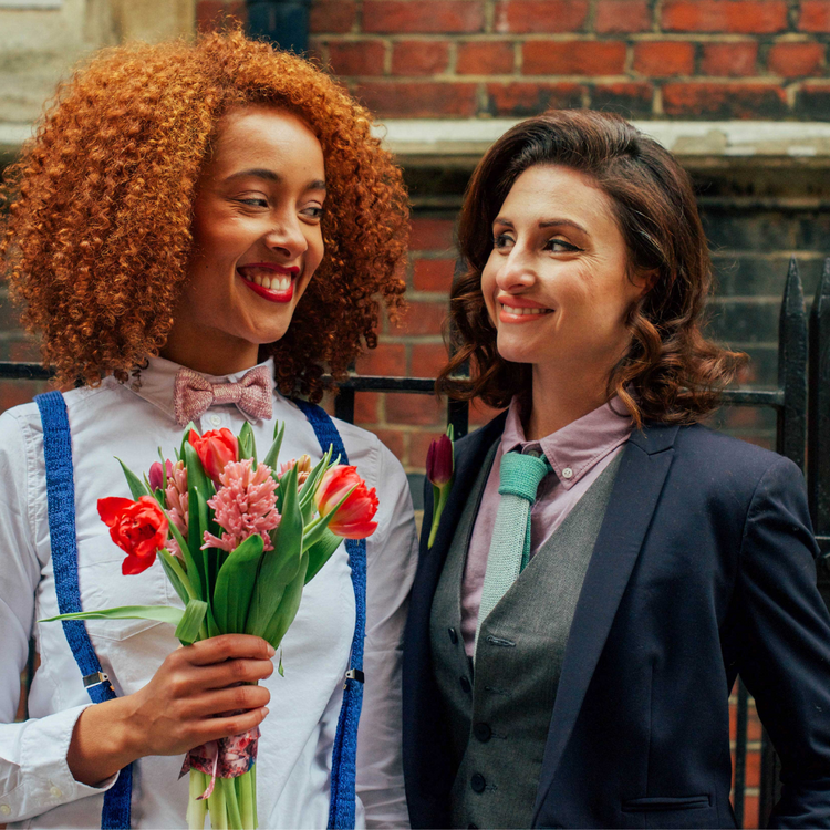 Two Women in wedding suits, suspenders, bow tie and tie, hold tulips and smile at each other.